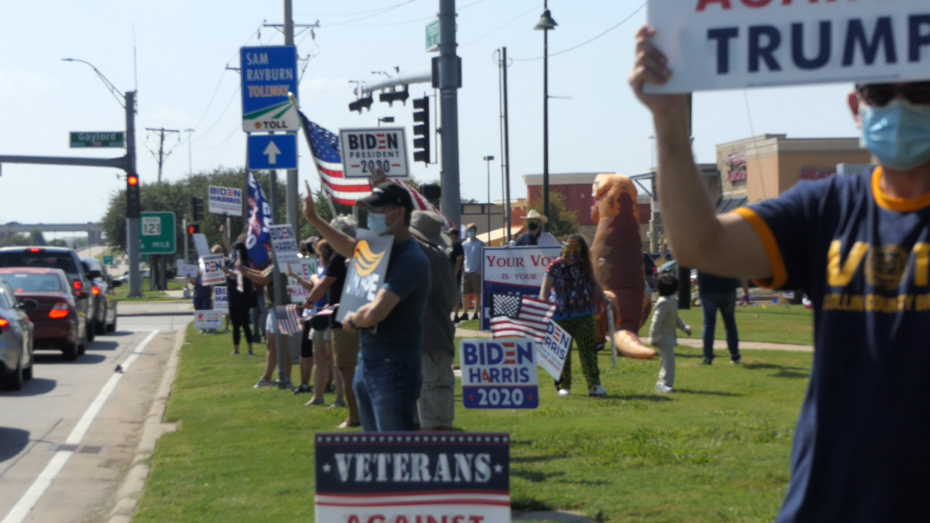 Biden-Harris supporters holding signs and flags on street curb, waving to cars